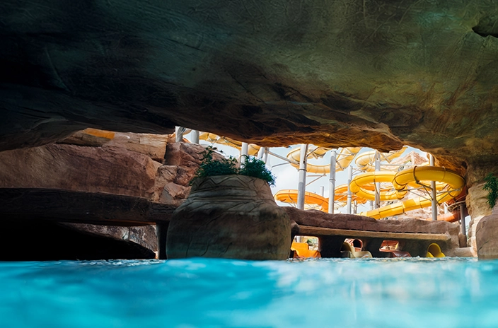 View from a cave-like area in a water park, with blue water in the foreground and bright yellow twisting slides in the background against rock formations.