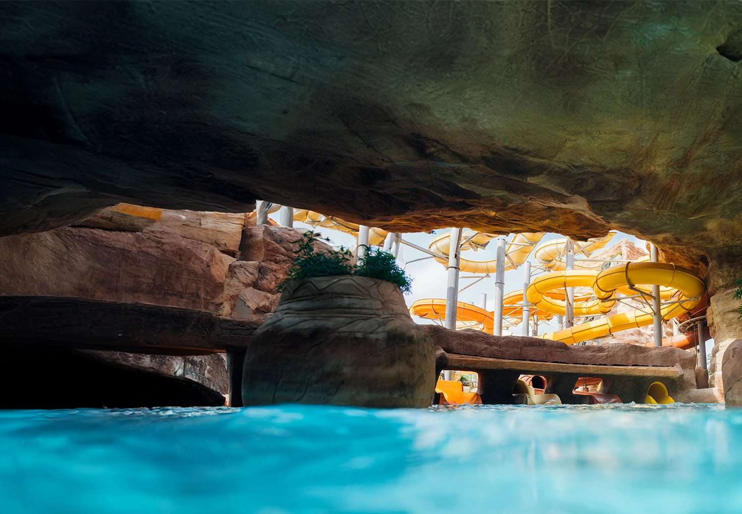 View from a cave-like area in a water park, with blue water in the foreground and bright yellow twisting slides in the background against rock formations.