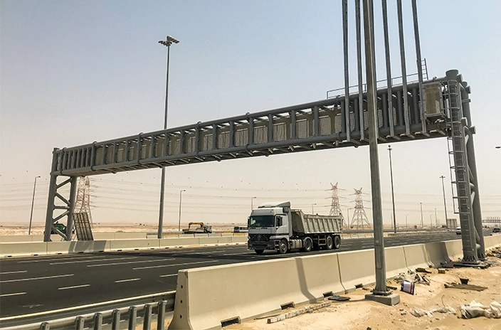 A truck on a highway under a large metal overhead structure in a desert setting.