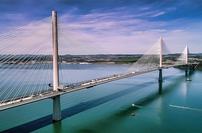 Queensferry Crossing bridge, showing its tall pylons and fan-like cable structure spanning across a calm, blue-green river on a clear day, with boats visible on the water below.