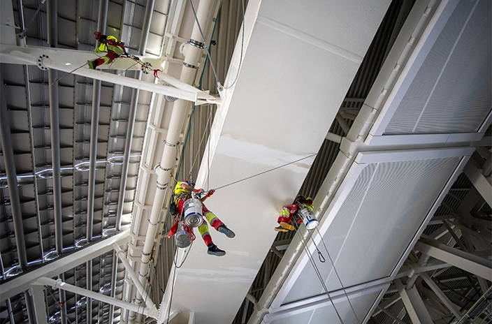 Three painters in safety gear are hanging on ropes high above the ground, applying paint to the beams and metal surfaces of the Santiago Bernabéu Stadium's roof.