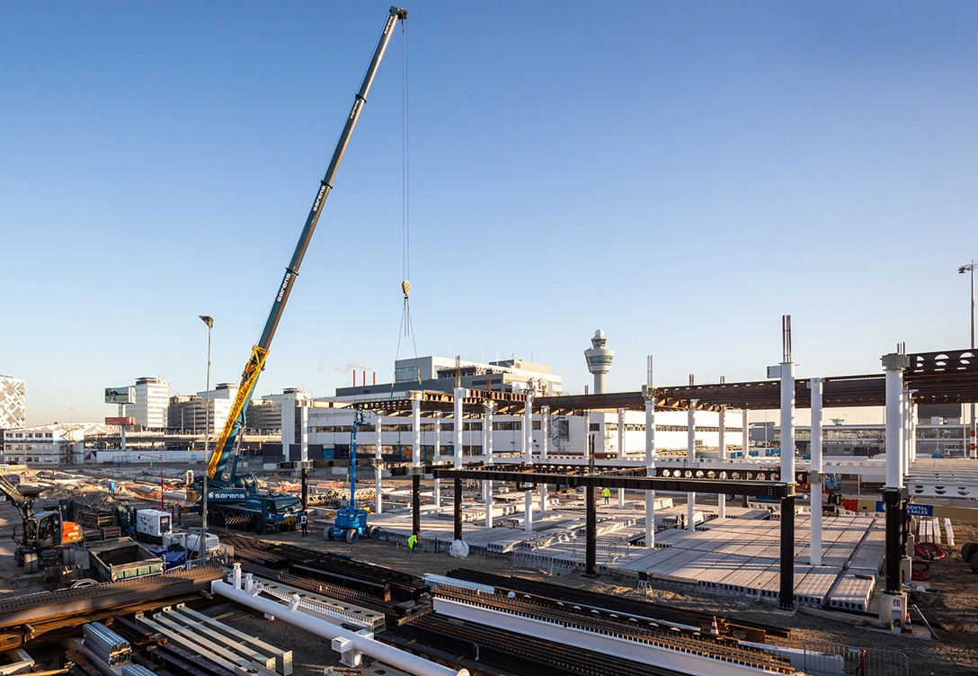 Construction site at Schiphol Airport with a large crane lifting materials, partially built structures, and various equipment.