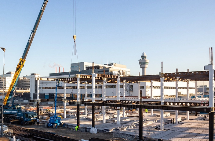 Construction site at Schiphol Airport with a large crane lifting materials, partially built structures, and various equipment.