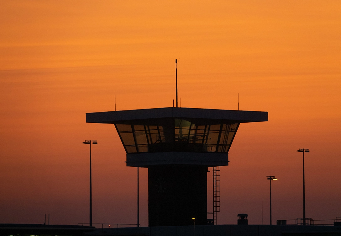 Silhouette of a Schiphol airport control tower against a vibrant orange sunset sky.