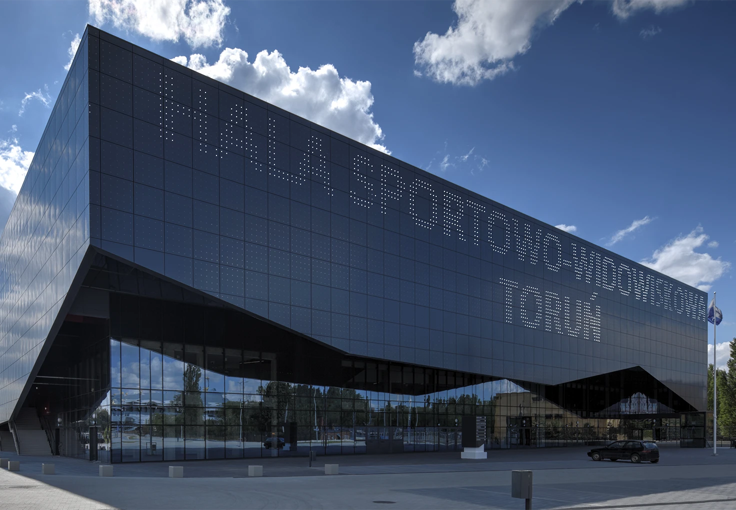 Modern glass-fronted sports and entertainment arena in Torun with the name “Hala Sportowo-Widowiskowa Toruń” displayed on the facade.