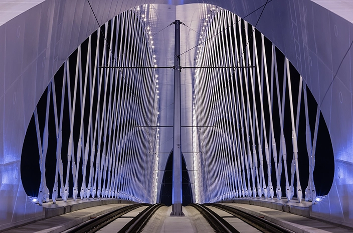 A nighttime view of Troja Bridge, illuminated by lights along its structure, with the symmetrical crisscrossing cables and railway tracks.