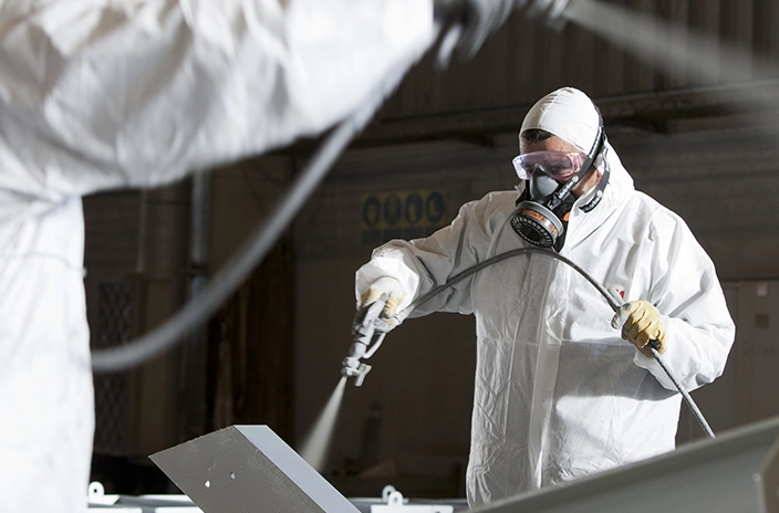 A worker in full protective gear, is spray painting a metal surface in an industrial setting.