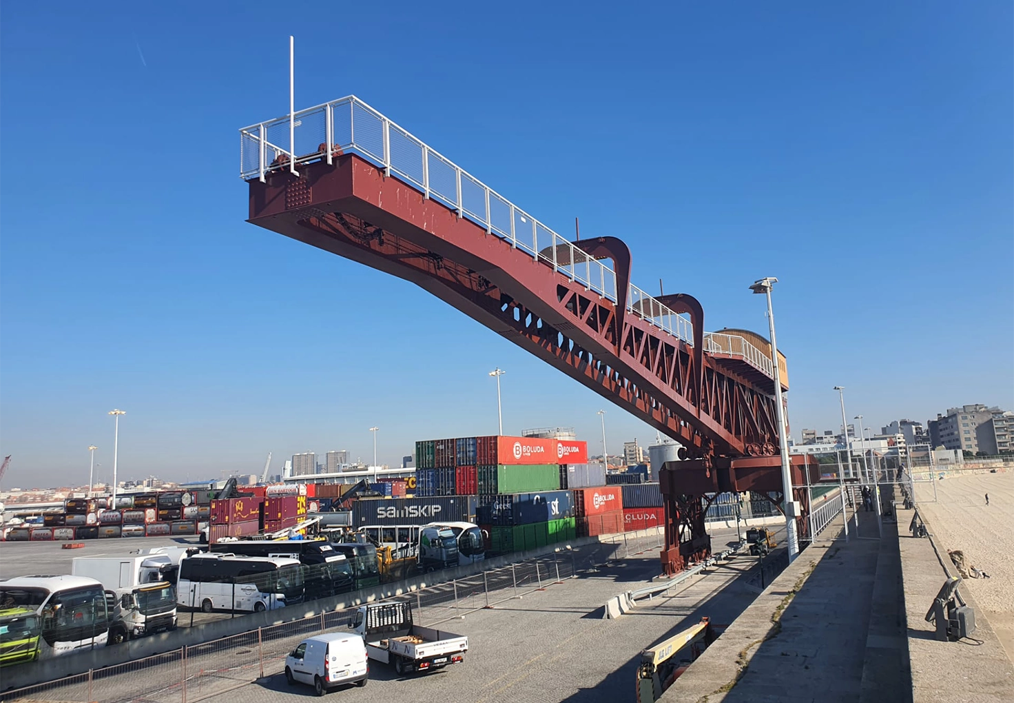 A large red Vesam port crane extends over a busy industrial area with trucks, containers, and equipment. Red port crane at Vesam spanning over trucks and shipping containers with cranes in the background under a clear sky.