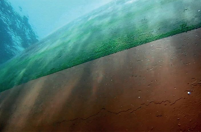 An underwater view of the Famenne's hull shows a clear contrast between algae-covered and clean areas, demonstrating Hempaguard's effectiveness after 23 months in fouling-aggressive waters.