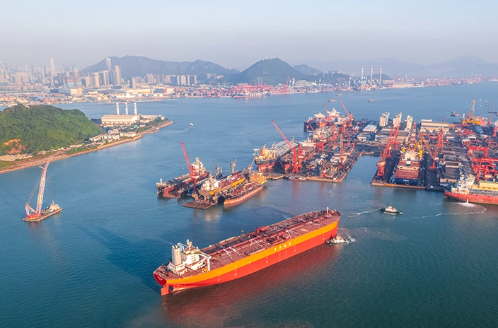 A large orange tanker from FPMC operator is seen moving through a busy industrial harbor with several ships, cranes, and facilities in the background.