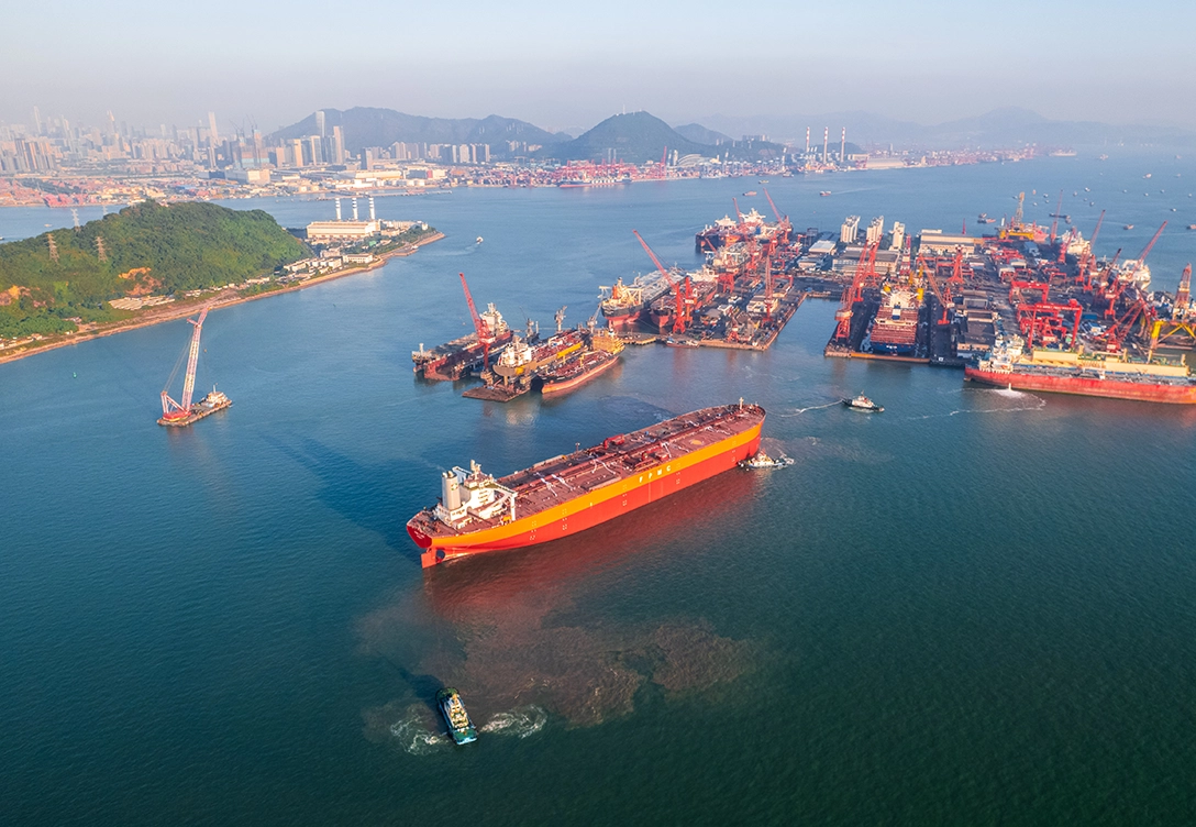 A large orange tanker from FPMC operator is seen moving through a busy industrial harbor with several ships, cranes, and facilities in the background.