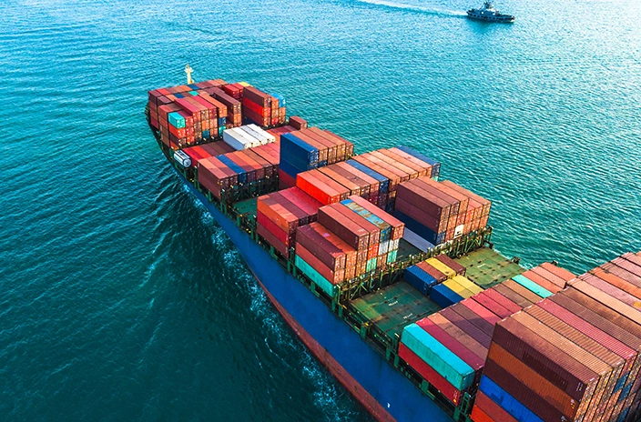 A neopanamax container vessel carrying colorful shipping containers sailing through calm waters, with a smaller ship visible in the distance.