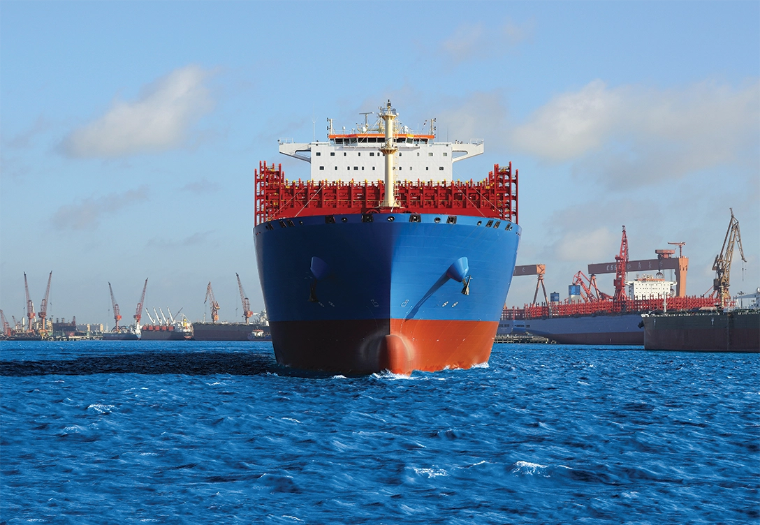 A large neopanamax container ship with a blue and red hull is shown from the front as it sails through a harbor, with cranes and other ships visible in the background.