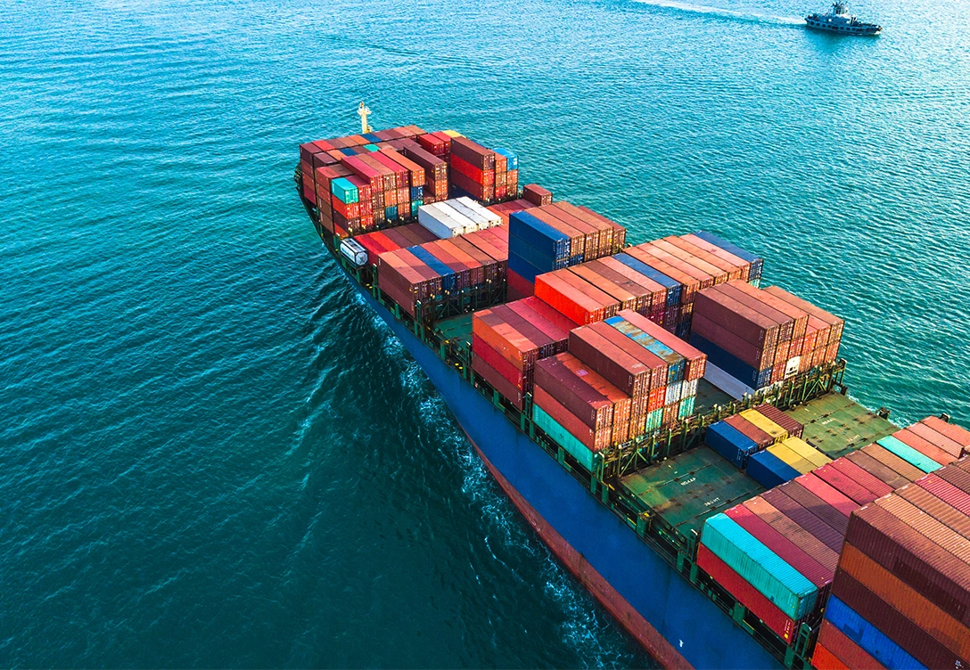 A neopanamax container vessel carrying colorful shipping containers sailing through calm waters, with a smaller ship visible in the distance.