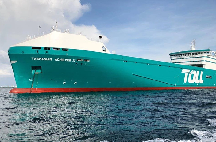 The Ro-Ro vessel "Tasmanian Achiever II" from Toll Shipping sailing in calm waters, featuring a teal and white hull with the company's logo displayed.