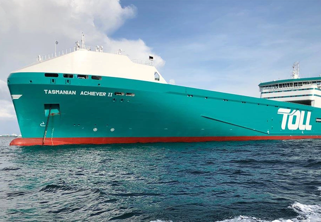 The Ro-Ro vessel "Tasmanian Achiever II" from Toll Shipping sailing in calm waters, featuring a teal and white hull with the company's logo displayed.