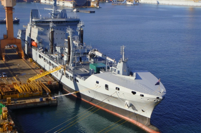 A UK Royal Navy vessel is docked at a shipyard, surrounded by equipment and cranes.