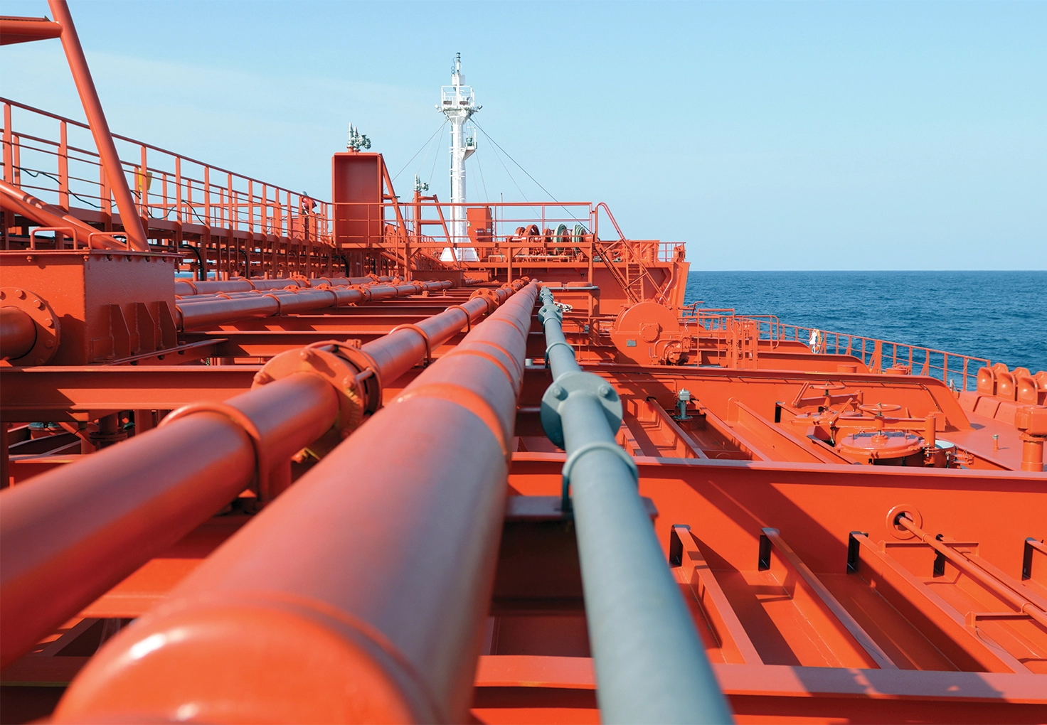 A view along the deck of a VLCC tanker shows a series of red pipelines and equipment stretching toward the bow, with the ocean and a clear sky in the background.