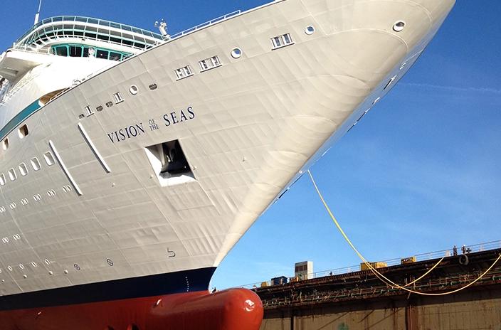 The bow of the ship "Vision of the Seas" from Royal Caribbean is shown in a shipyard, with it's white hull and the ship's name clearly visible on the side.