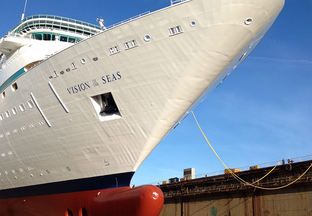 The bow of the ship "Vision of the Seas" from Royal Caribbean is shown in a shipyard, with it's white hull and the ship's name clearly visible on the side.