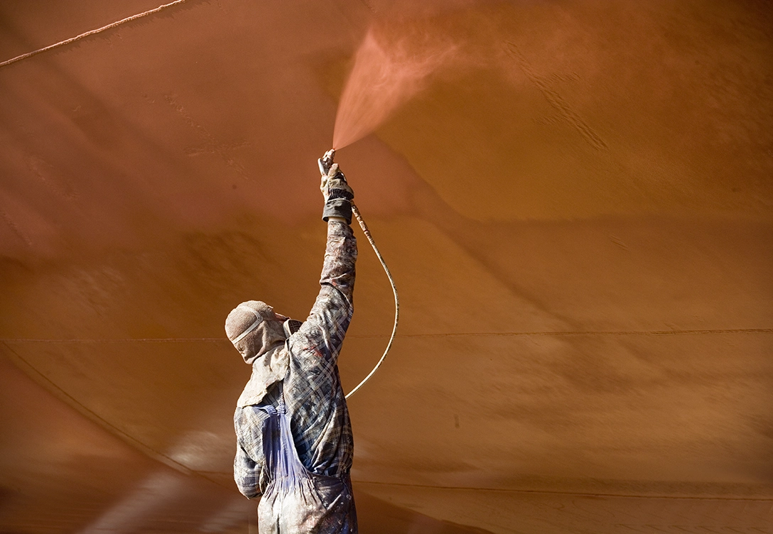 A worker wearing protective gear uses a spray gun to coat a vessel with red paint or coating material.