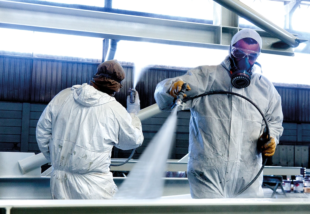 Two workers in protective suits and masks spray painting a metal structure.