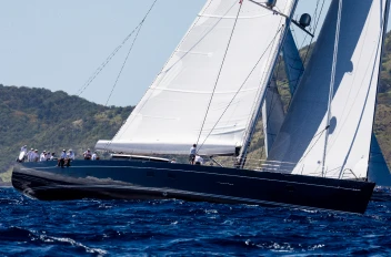 Superyacht G2 sailing on deep blue waters, with its white sails fully raised and crew members visible on deck, against a backdrop of hills.