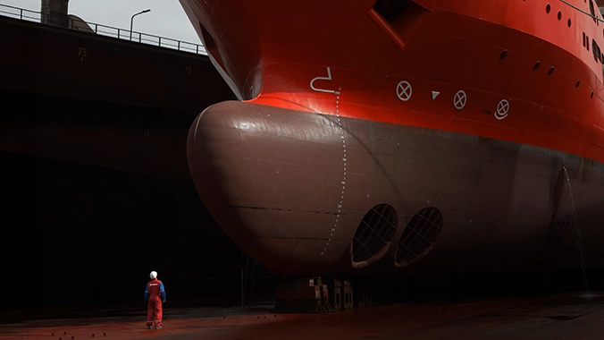 A worker in red coveralls and a white helmet stands in a dry dock, looking up at the massive bow of a red and brown ship undergoing maintenance.