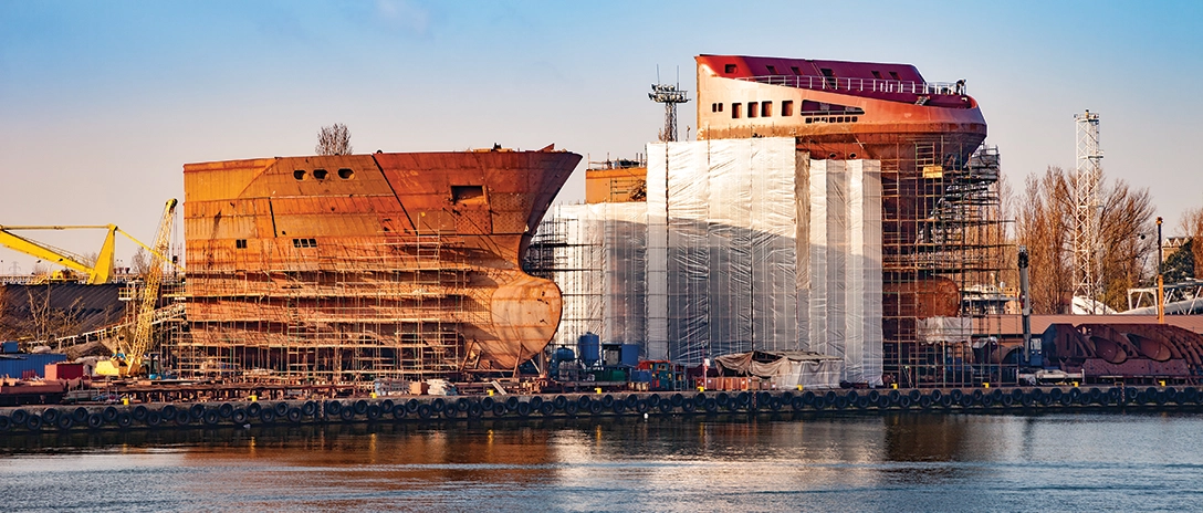 A newbuild ship under construction, with scaffolding surrounding its hull, sits at a shipyard by the water, partially covered with protective sheets.