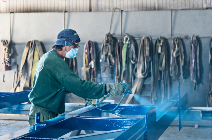 A worker wearing protective gear applies a blue coating to a metal structure using a spray gun in an industrial setting.