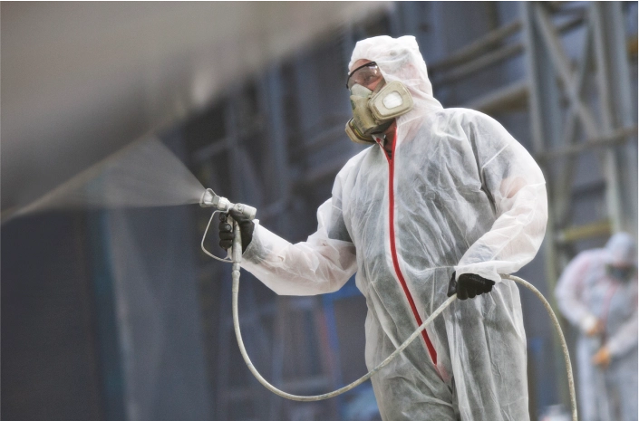 Detailed view of a worker in a full protective suit and mask applies industrial coating with a spray gun in a large facility.
