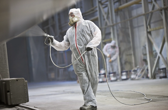 A worker in a full protective suit and mask applies industrial coating with a spray gun in a large facility.