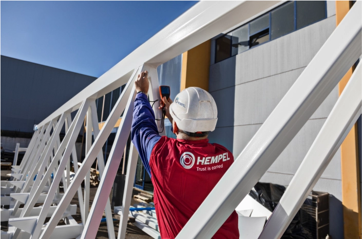 A coating advisor from Hempel inspecting the surface of a large white metal structure with a measuring device.