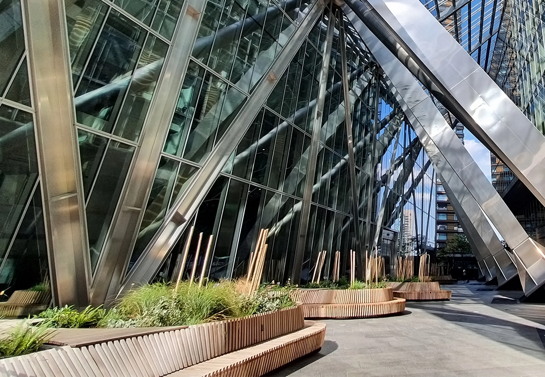 The exterior of a modern glass and metal building with angled supports and landscaped benches outside.