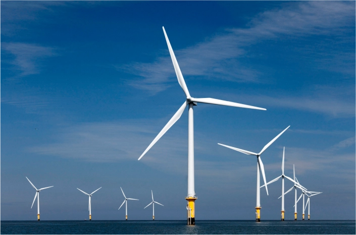A row of offshore wind turbines against a clear blue sky, standing in calm waters.