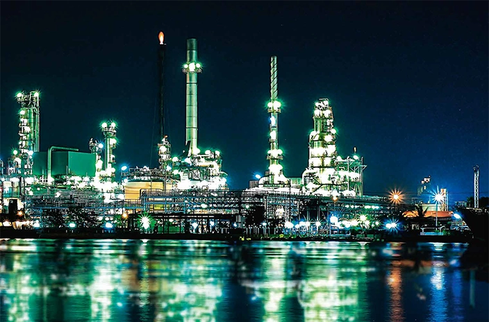 A brightly lit Elco's oil and gas facility at night, with reflections on the water.