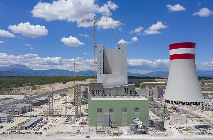 A large construction site featuring the Ptolemais Unit V power plant with a tall red and white cooling tower.