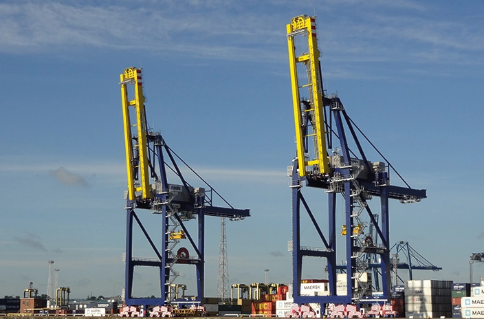 Two large yellow and blue Paceco cranes from the side at a shipping port, positioned over stacks of cargo containers.