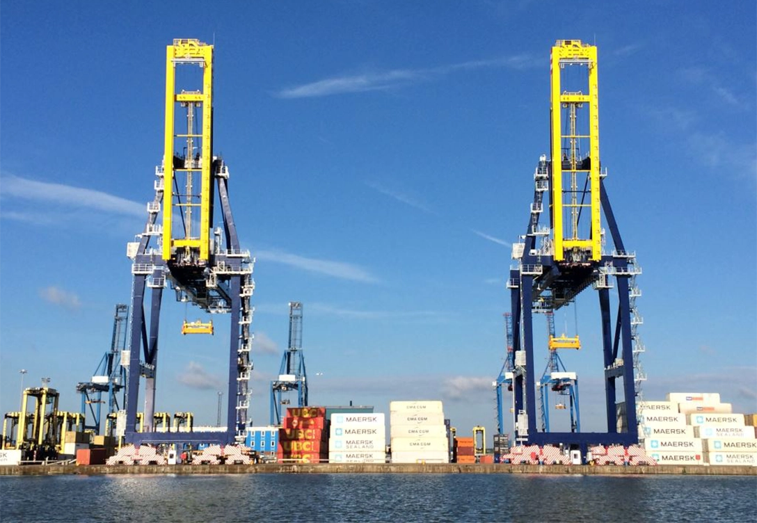 Two large yellow and blue Paceco cranes at a shipping port, positioned over stacks of cargo containers near the water.