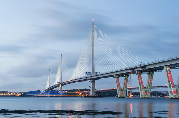 Queensferry Crossing bridge with tall pylons and illuminated cables at dusk, a blurred vessel passing underneath, and reflections on calm water under a cloudy sky.