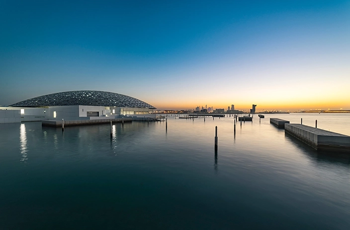 A view of the Louvre Abu Dhabi Museum, with its domed roof, sitting on the water as the sun sets in the background