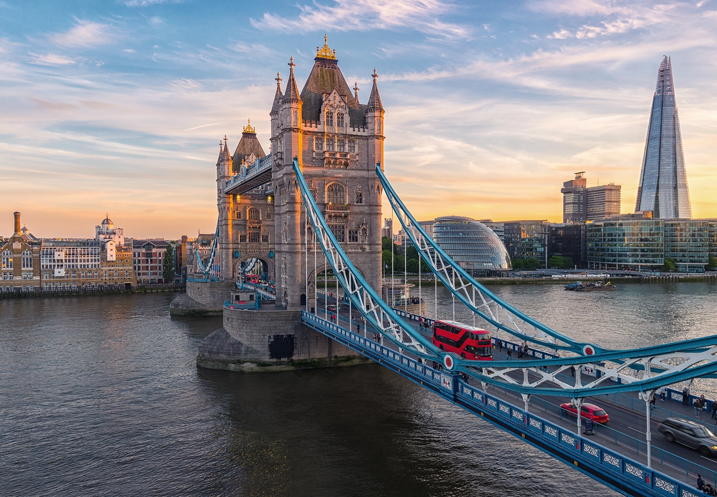 Tower Bridge in London at sunset, with a red double-decker bus crossing and the Shard in the background along the River Thames.
