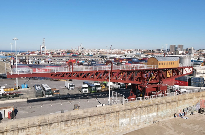 Red port crane at Vesam spanning over trucks and shipping containers with cranes in the background under a clear sky.