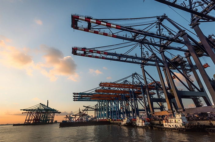 ZPMC port cranes at sunset, towering over docked ships and barges, with the sky and water reflecting the warm glow of the setting sun.
