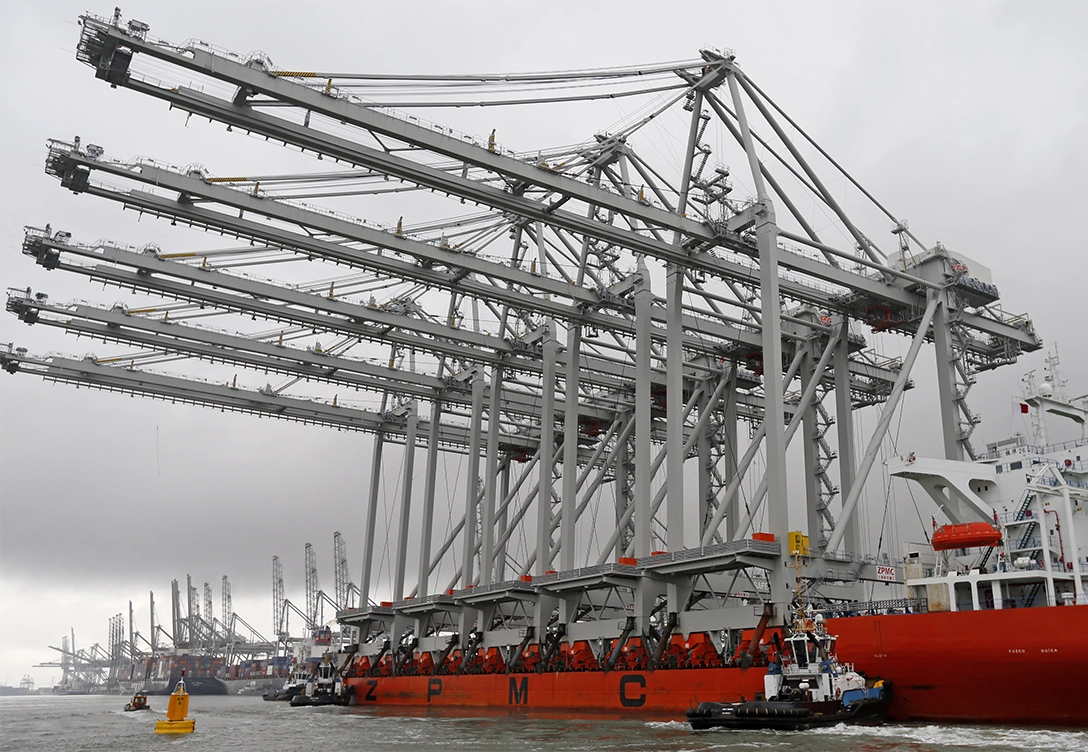 ZPMC port cranes on a barge, towering over the water with a tugboat alongside, under a cloudy sky.