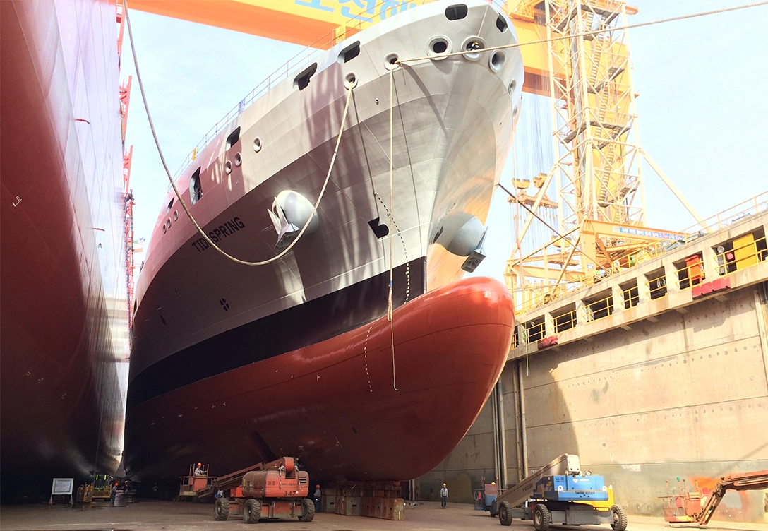 UK Navy vessel in a dry dock, with its gray and red hull towering above, surrounded by equipment and workers in the shipyard.