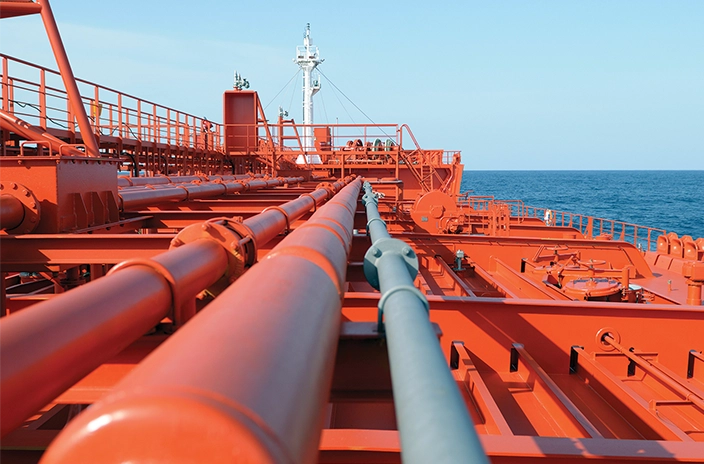A view along the deck of a VLCC tanker shows a series of red pipelines and equipment stretching toward the bow, with the ocean and a clear sky in the background.